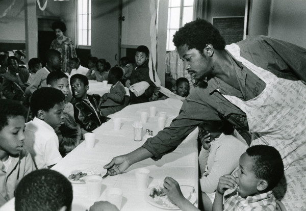 Charles Bursey hands plate of food to a child seated at Free Breakfast Program. Photo courtesy of Pirkle Jones and Ruth-Marion Baruch.